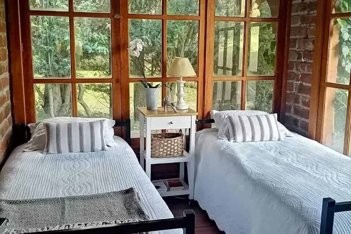 Quarto com duas camas, uma mesa de cabeceira branca, abajur e janelas amplas com vista para o verde.