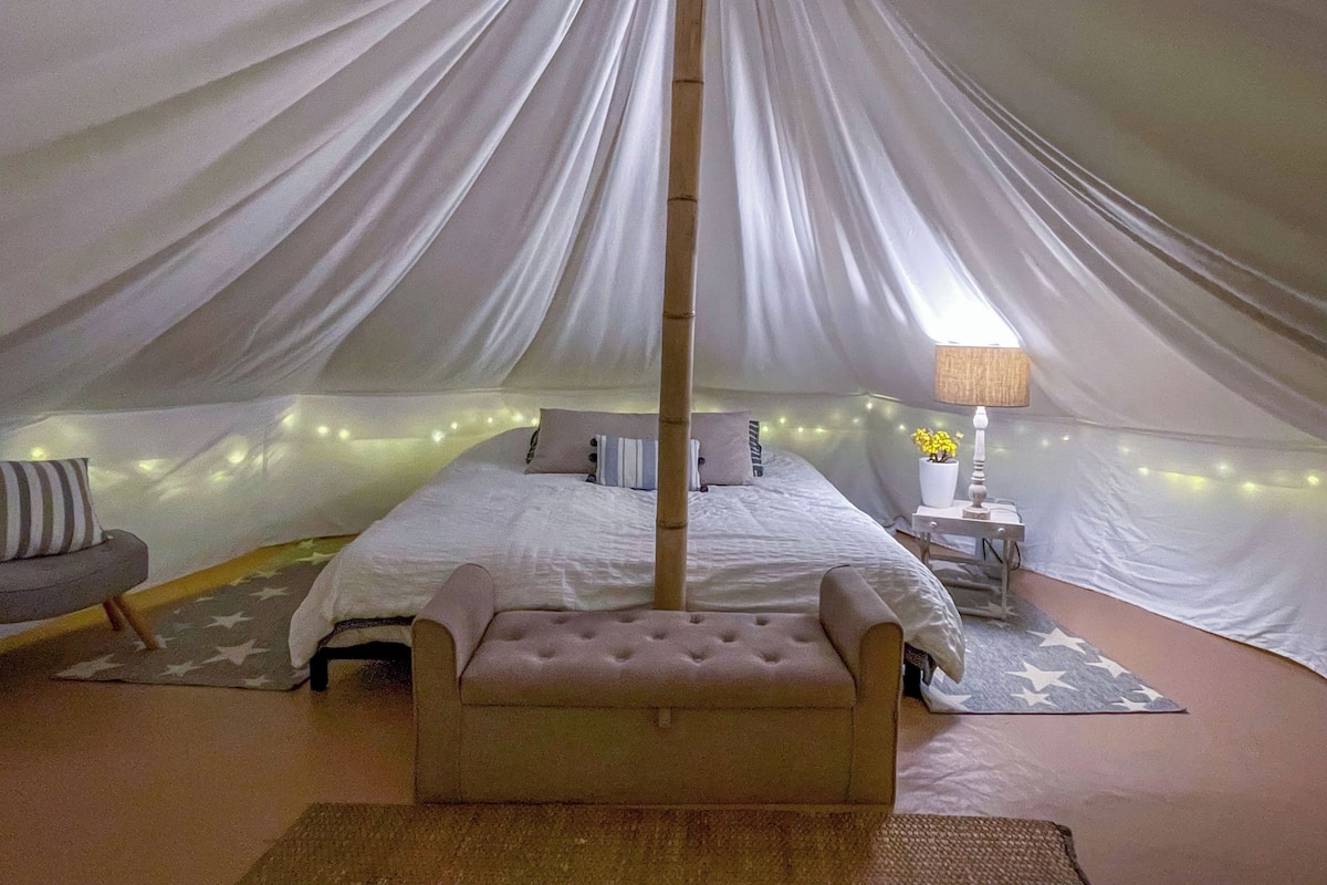 Um quarto com tenda, com uma cama, mesa de cabeceira, abajur e um banco.