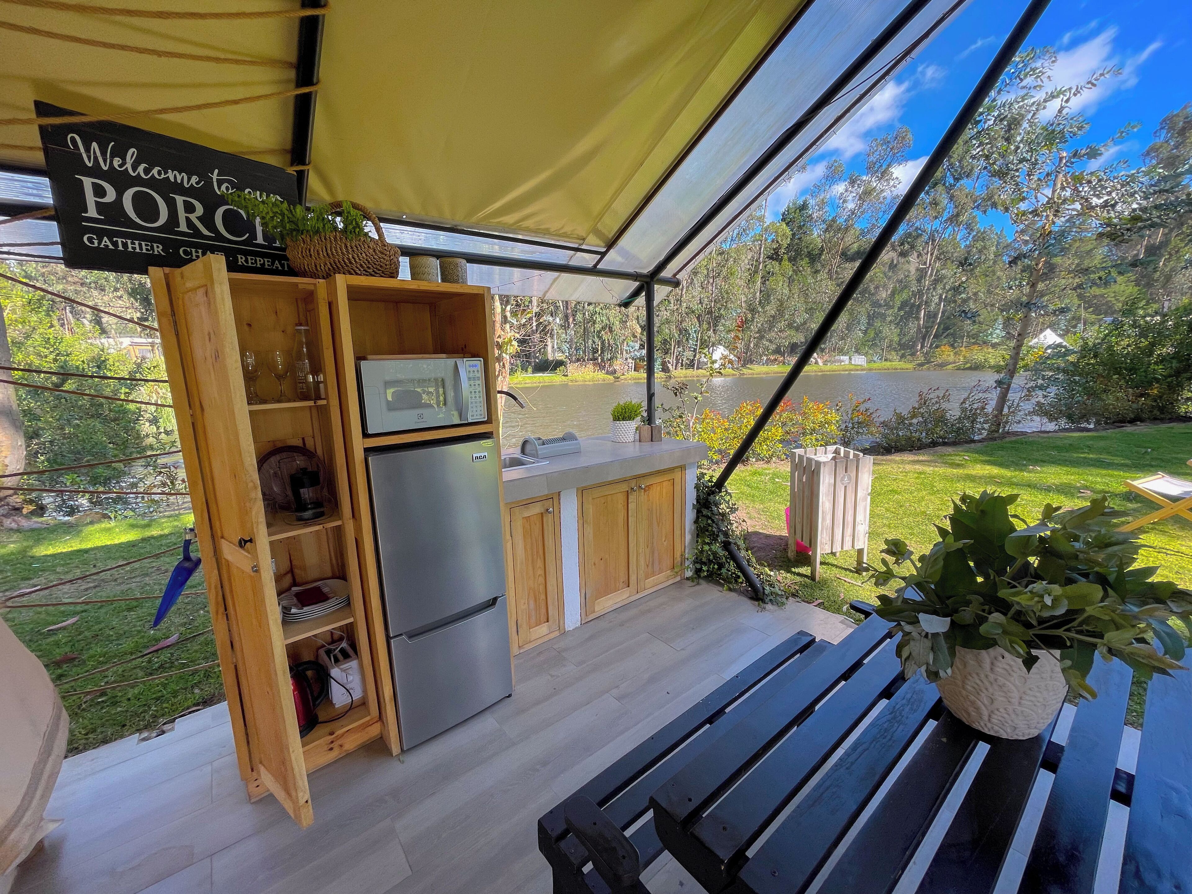 An outdoor kitchen area with a stainless steel refrigerator, wooden cabinets, and a microwave. There is a sign that reads "Welcome to your PORCH GATHER CHART REPEAT." A bench and a potted plant are also visible.