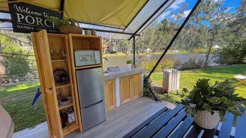 An outdoor kitchen area with a stainless steel refrigerator, wooden cabinets, and a microwave. There is a sign that reads "Welcome to your PORCH GATHER CHART REPEAT." A bench and a potted plant are also visible.