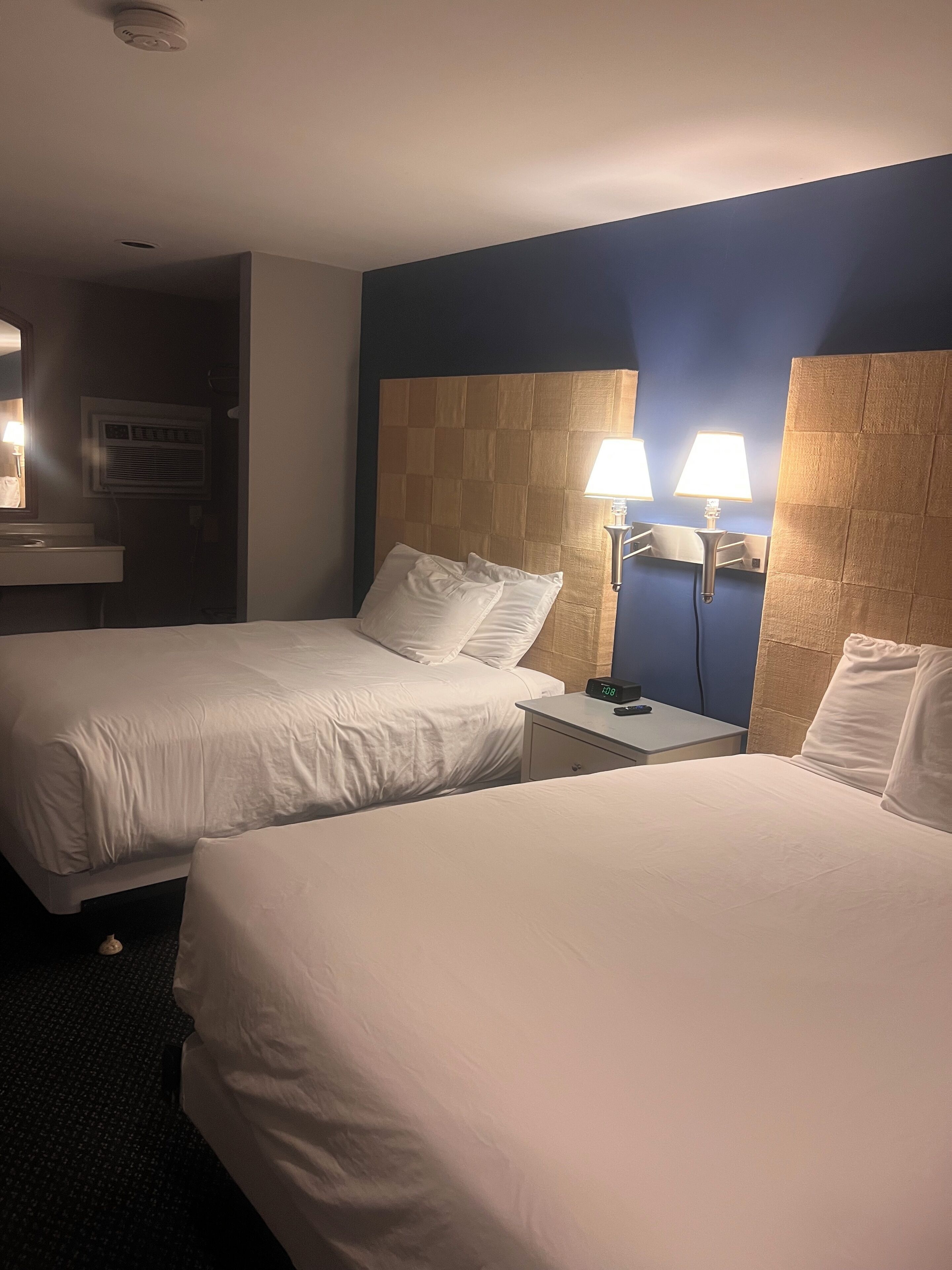 Une chambre d’hôtel avec deux lits, une table de chevet avec une horloge, une lampe fixée au mur, et une salle de bain visible au fond.