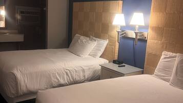 Une chambre d’hôtel avec deux lits, une table de chevet avec une horloge, une lampe fixée au mur, et une salle de bain visible au fond.