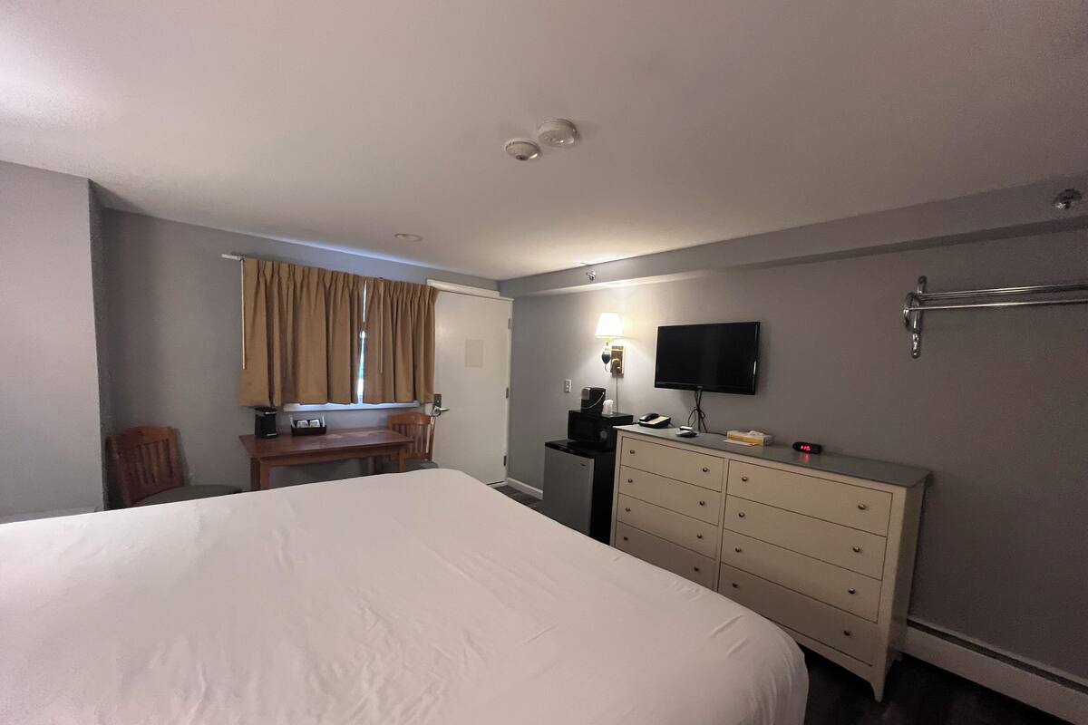 Ein Hotelzimmer mit einem großen Bett, einem Schreibtisch mit Stuhl, einem Schminktisch und einem an der Wand befestigten Fernseher sowie einem Fenster mit Vorhängen.