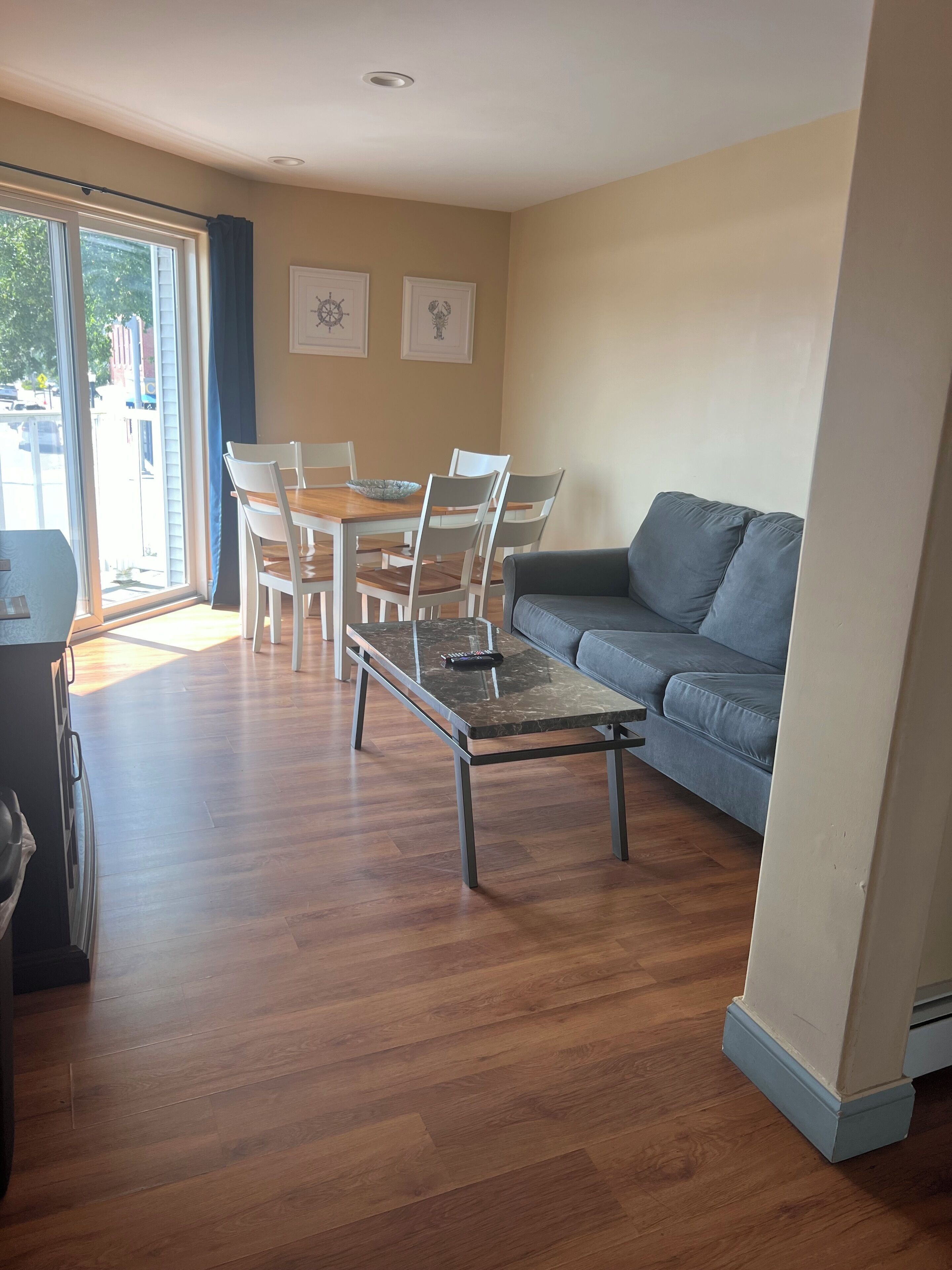 A living room with a grey sofa, a wooden dining table with chairs, and a glass door leading outside.