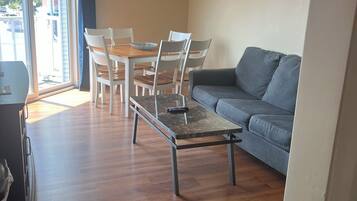 A living room with a grey sofa, a wooden dining table with chairs, and a glass door leading outside.