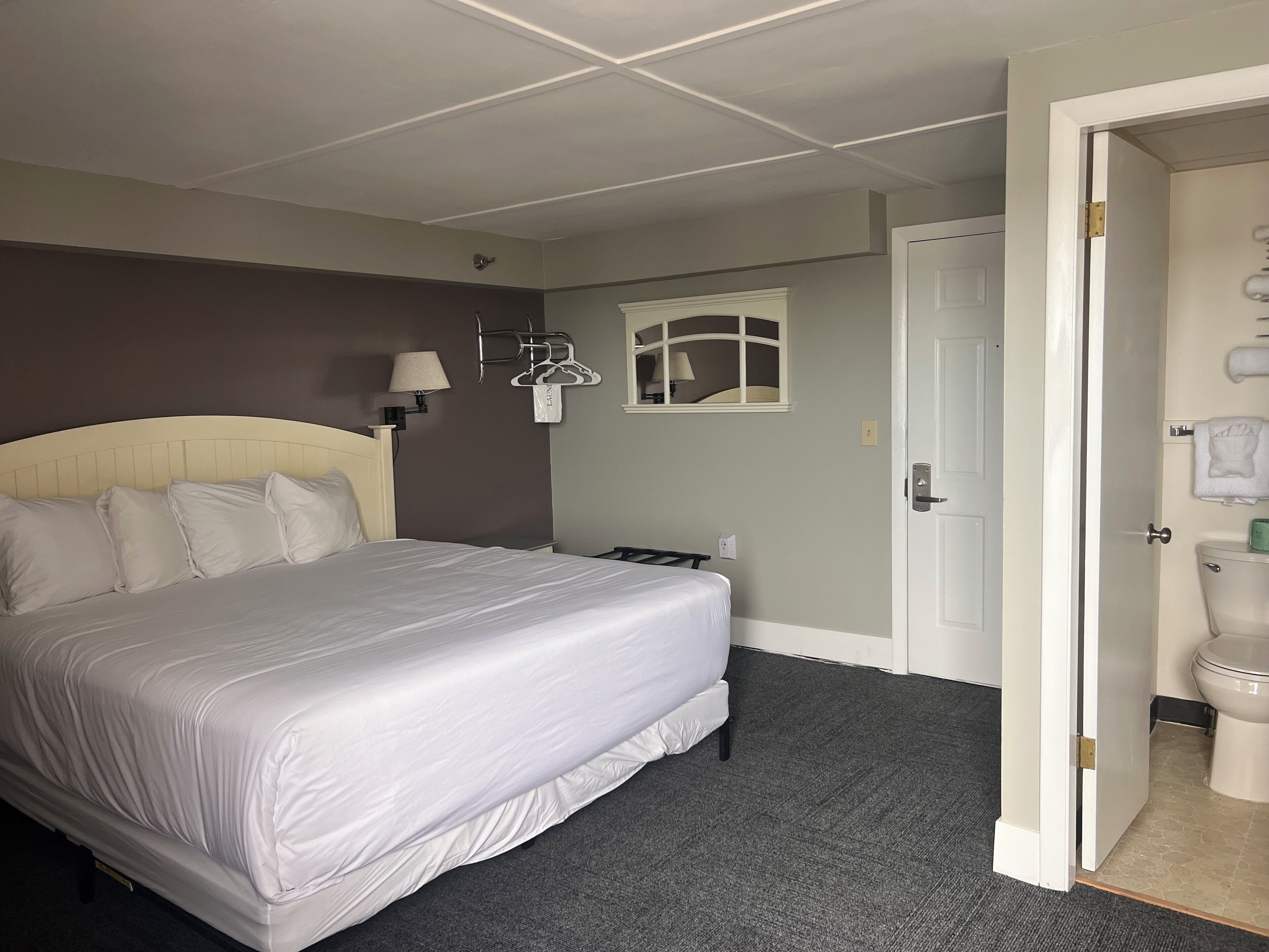 A hotel room with a large bed, a nightstand, a wall-mounted lamp, a window with a view, and a door leading to the bathroom.