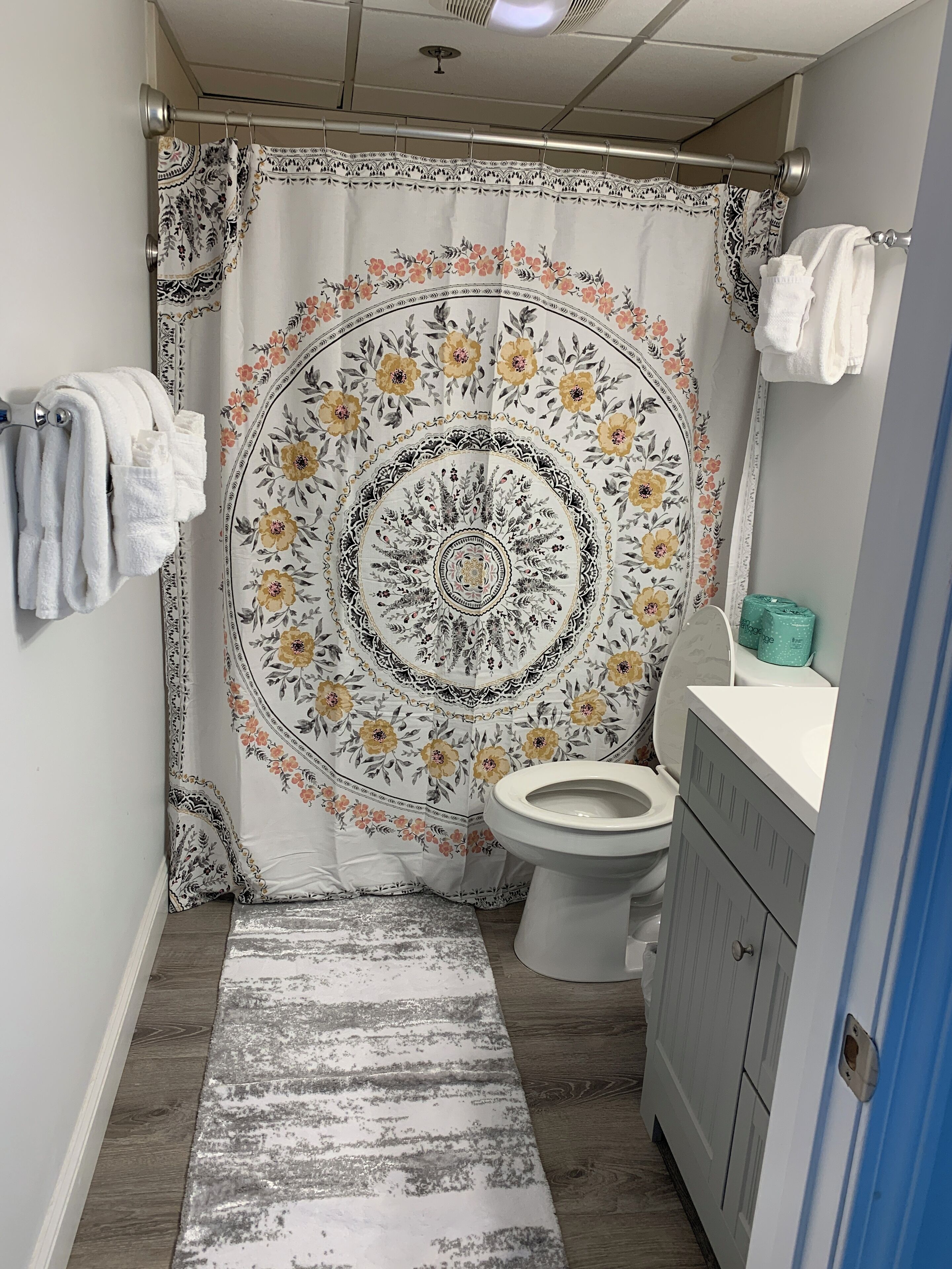 Une salle de bain avec un rideau de douche orné d’un motif floral, des toilettes, un lavabo avec un miroir et un porte-serviettes avec des serviettes.