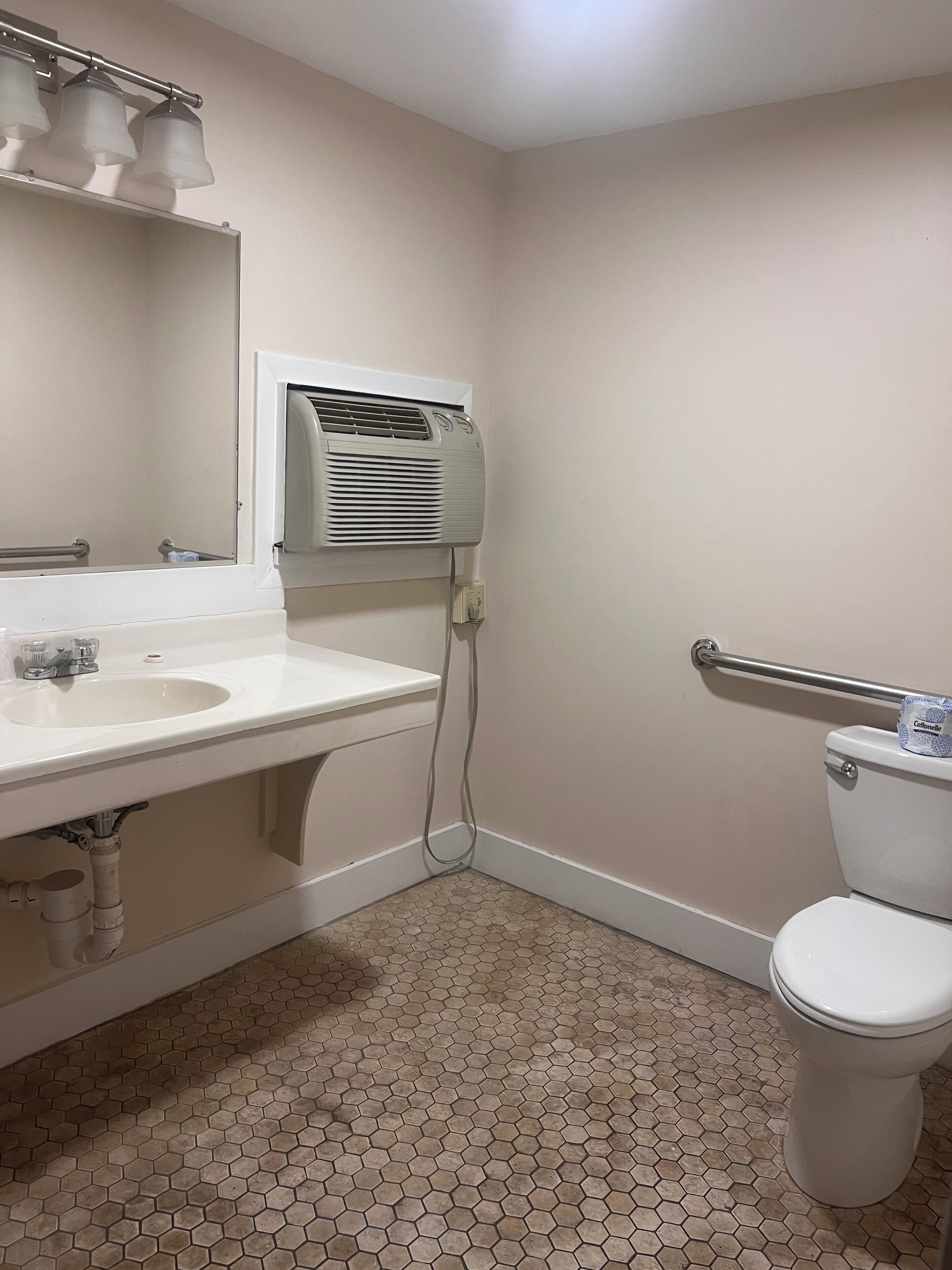 Een badkamer met een witte wastafel, spiegel en toilet. Er is een plafondventilator en een handgreep in de buurt van het toilet.