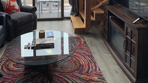 A modern living room with a glass coffee table, a sofa with a colorful throw, a TV on a wooden cabinet, and a view of the outdoors through large windows. - Trade Winds on the Bay (Rockland)