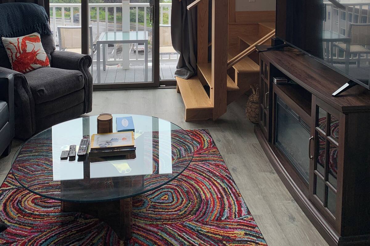 Ein modernes Wohnzimmer mit einem Glastisch, einem Sofa mit farbenfroher Decke, einem Fernseher auf einer Holzkommode und Blick ins Freie durch große Fenster.