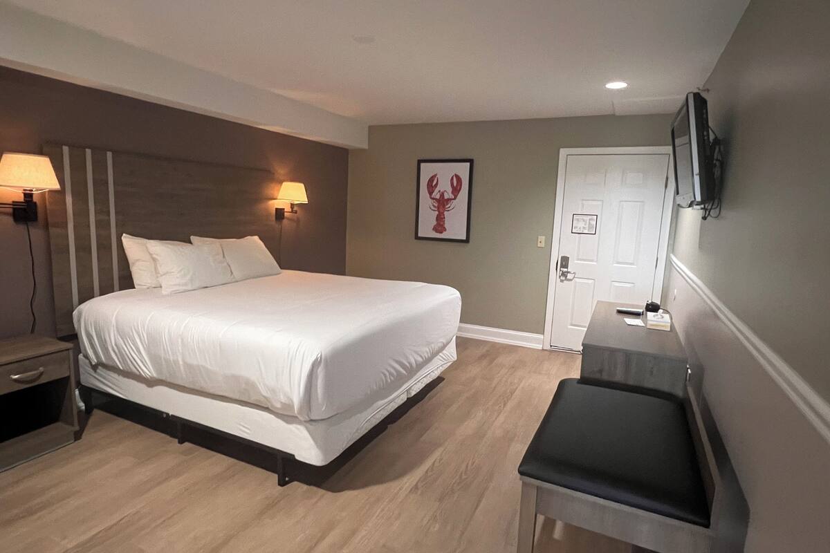 Ein Hotelzimmer mit einem großen Bett, Nachttischlampen, einem Schreibtisch mit Stuhl, einer Tür und einem an der Wand befestigten Fernseher.