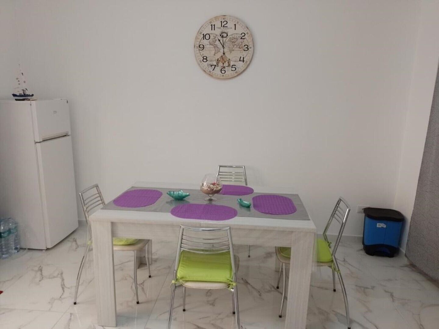 Un coin repas avec un sol en marbre, une table blanche, quatre chaises dotées de coussins verts et une horloge murale.