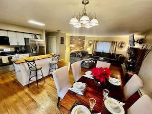 A modern kitchen with a central island, a dining area with a wooden table set for a meal, and a living room with a sofa and a fireplace.