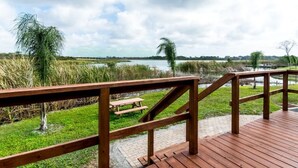 A wooden deck with a picnic table and benches overlooking a grassy area with reeds and a body of water.