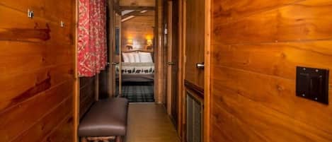 A narrow wooden hallway with a bench, a bed with a plaid bedspread, and a red curtain.
