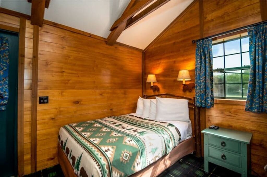 Habitació de cabana de fusta amb un llit, dues làmpades de nit, una finestra amb cortines i una petita taula de nit.