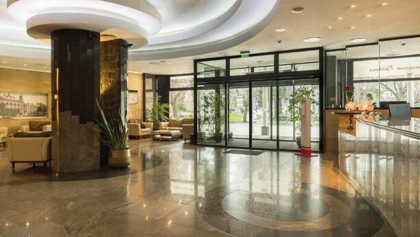 Un hall espacioso con diseño moderno, que presenta una amplia columna central, zonas de estar confortables y una entrada de cristal hacia el exterior.