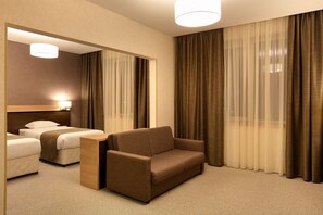 A hotel room with a sofa, two beds, a mirror, and a large window with curtains. - Danube Plaza Hotel (Ruse)