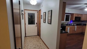 A hallway leading to a kitchen with wooden flooring, a door with a window, and framed pictures on the wall.