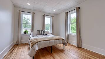 A bedroom with a bed, wooden flooring, and large windows with curtains.