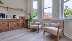 A modern kitchen with wooden cabinets, a coffee machine, and a plant. There are two beige chairs and large windows with a view of a brick building.
