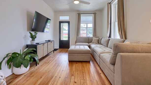 A living room with a sectional sofa, a flat-screen TV mounted on the wall, a ceiling fan, and a large window with curtains.