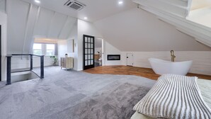 A spacious attic bedroom with a large window, a white bathtub, and a wooden floor.