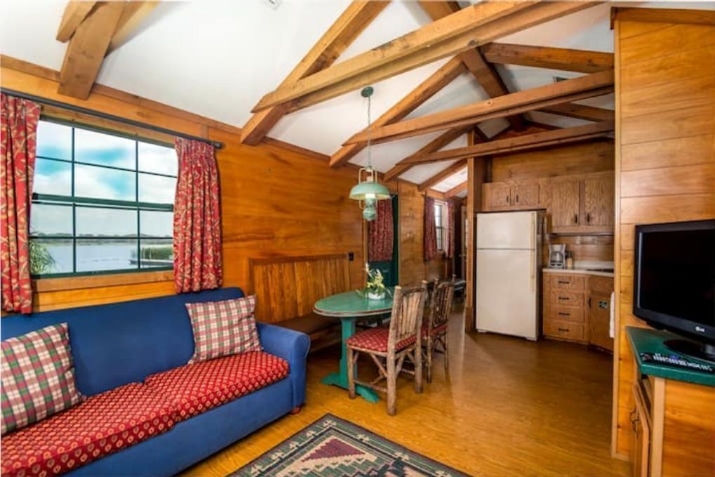 Ein gemütliches Blockhaus-Interieur mit Holzbalkendecke, einem blauen Sofa mit gemusterten Kissen, einem Esstisch mit Stühlen und einer Küchenzeile mit Schränken und einem Kühlschrank.