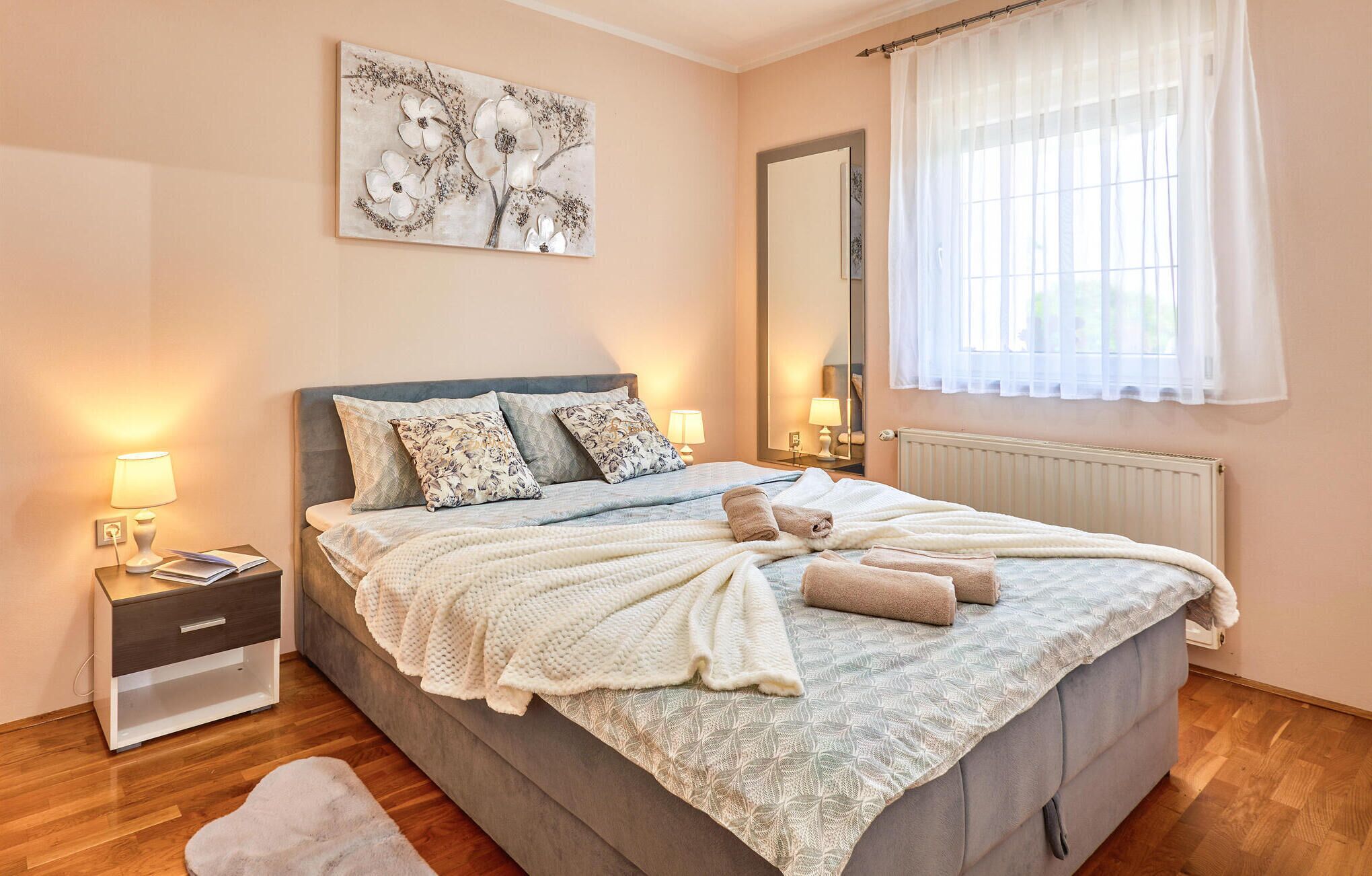 A bedroom with a bed, bedside lamps, a nightstand, a mirror, and a window with curtains.