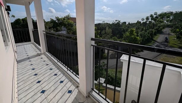 A balcony with a tiled floor and a metal railing, overlooking a residential area with trees and a road. - Hotel Arda Bali (Negara)