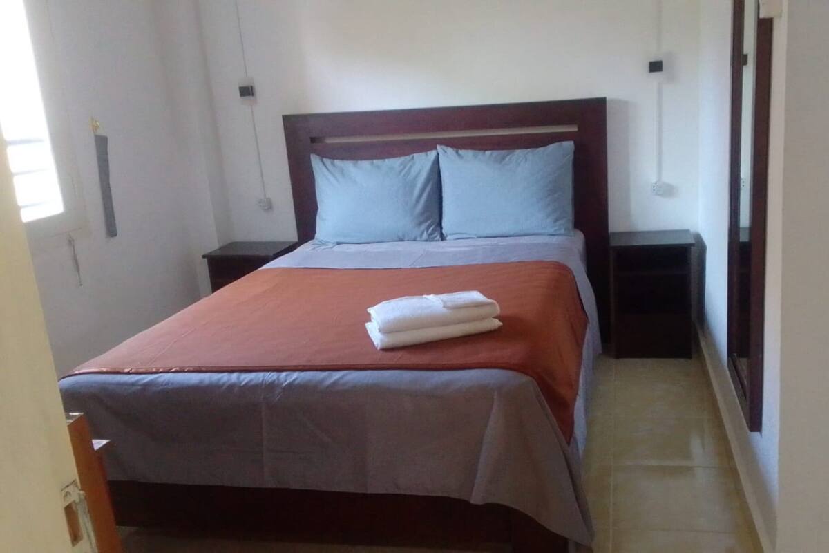 A neatly made bed with a red and grey bedspread, two pillows, and a folded white towel. There are two bedside tables with lamps, and the room has a simple, clean design.