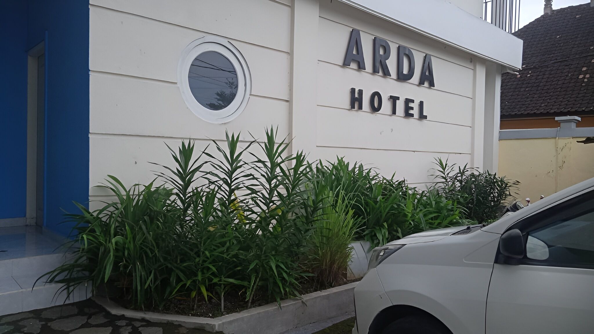 Un edificio con un letrero que dice "ARDA HOTEL". Tiene una ventana circular en la pared y un coche blanco estacionado en la parte delantera.