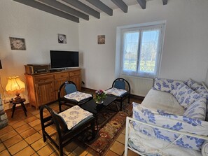 A living room with a TV, wooden cabinet, two chairs, a table, a sofa, and a window.