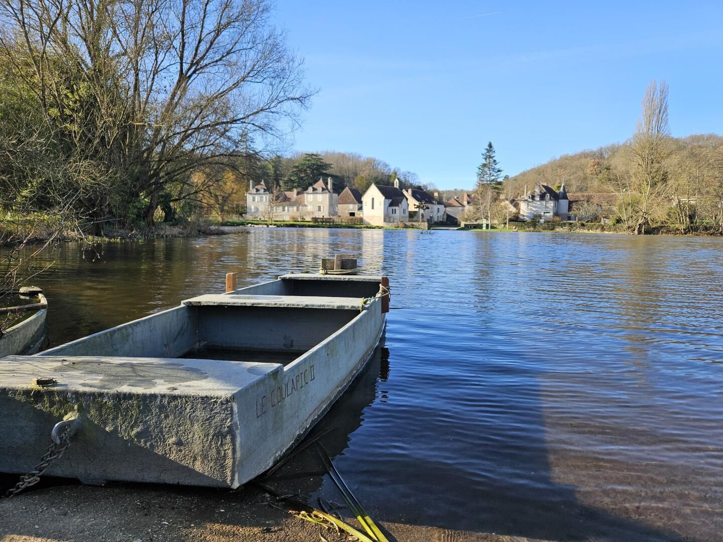 A calm river with boats docked, a clear sky, and houses in the background.