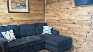 Une pièce chaleureuse avec un canapé d’angle bleu foncé, des murs en bois, un tableau représentant un paysage encadré et un plancher en bois.
