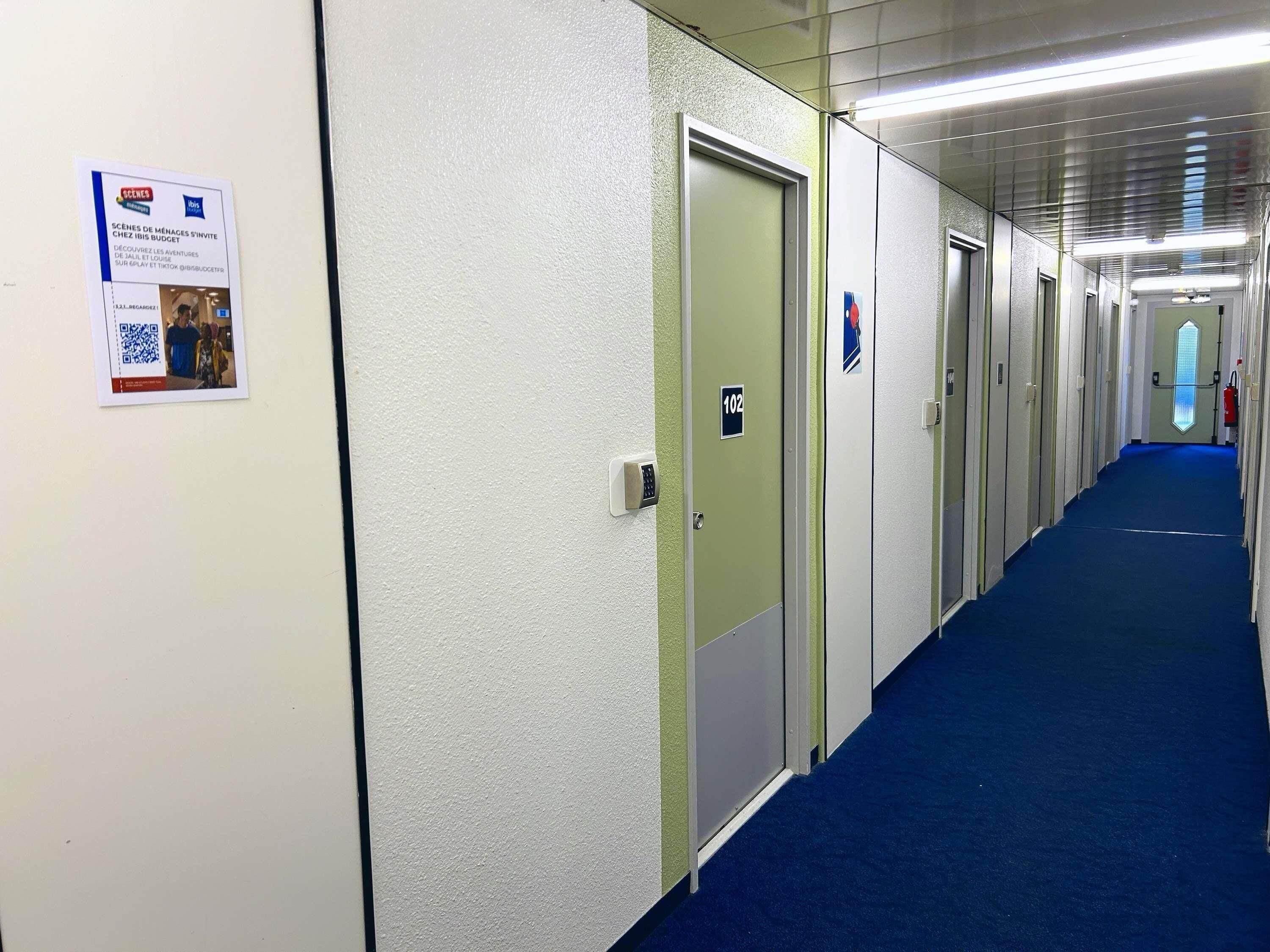 Pasillo de hotel con puertas numeradas 102, alfombra azul y un cartel en la pared.