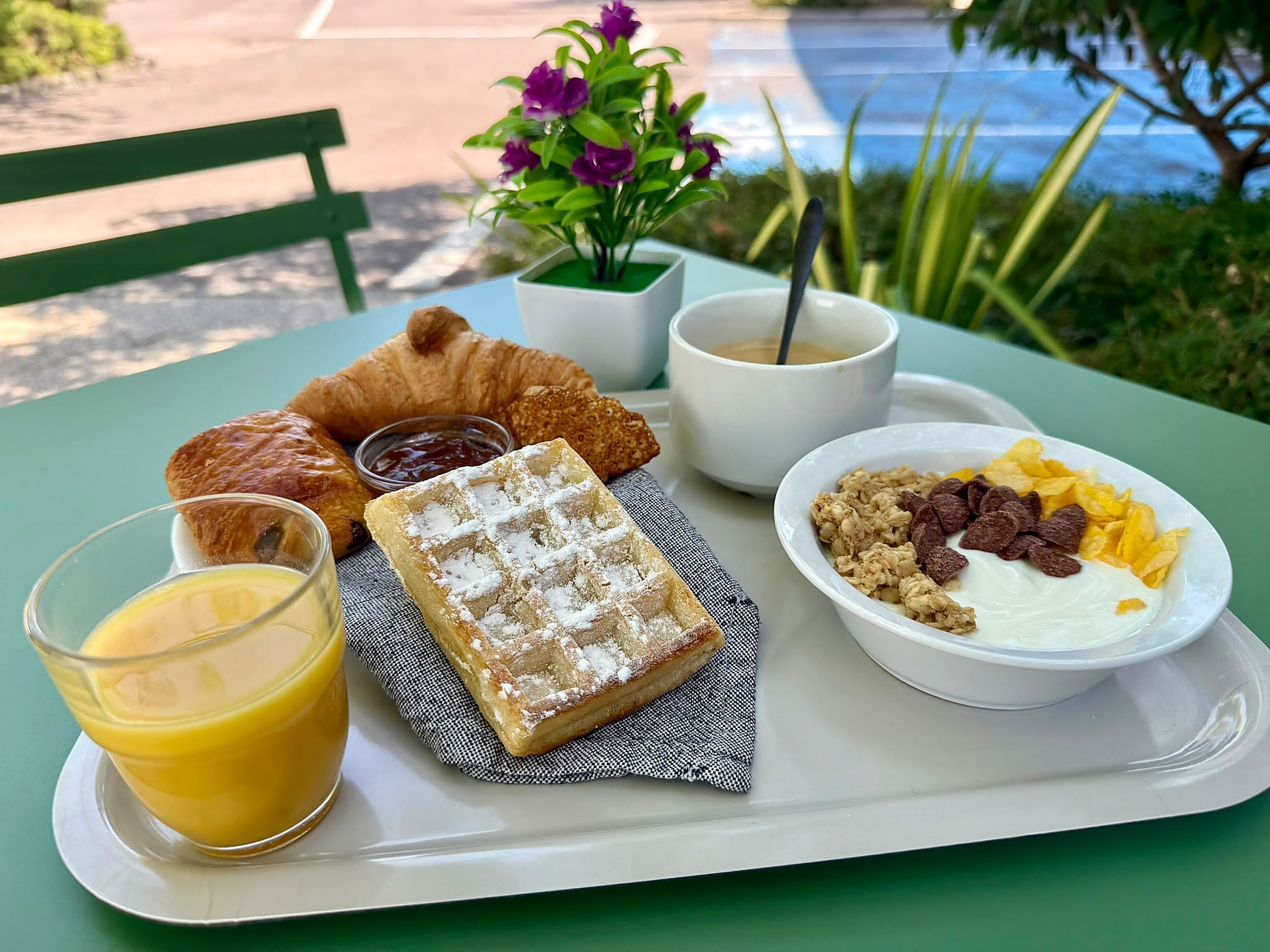 A breakfast tray with croissants, a waffle, a bowl of cereal with fruits, a glass of orange juice, and a cup of coffee.