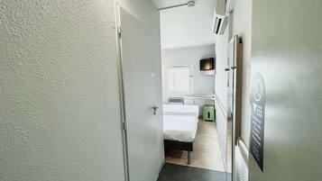 A narrow hallway leading to a bedroom with a bed, a small green stool, a window, and a mounted TV.