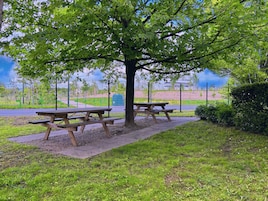 Una mesa de picnic bajo un árbol en un parque, con un parque infantil al fondo.