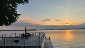A wooden deck with outdoor seating overlooking a calm body of water during sunset. - Beautifully updated home on the bay and golf course with dock and raised deck! (Sodus Point)