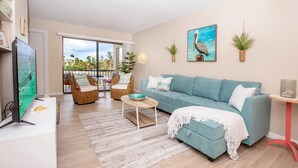 A modern living room with a teal sofa, a flat-screen TV, wicker chairs, and a balcony with a view of palm trees.