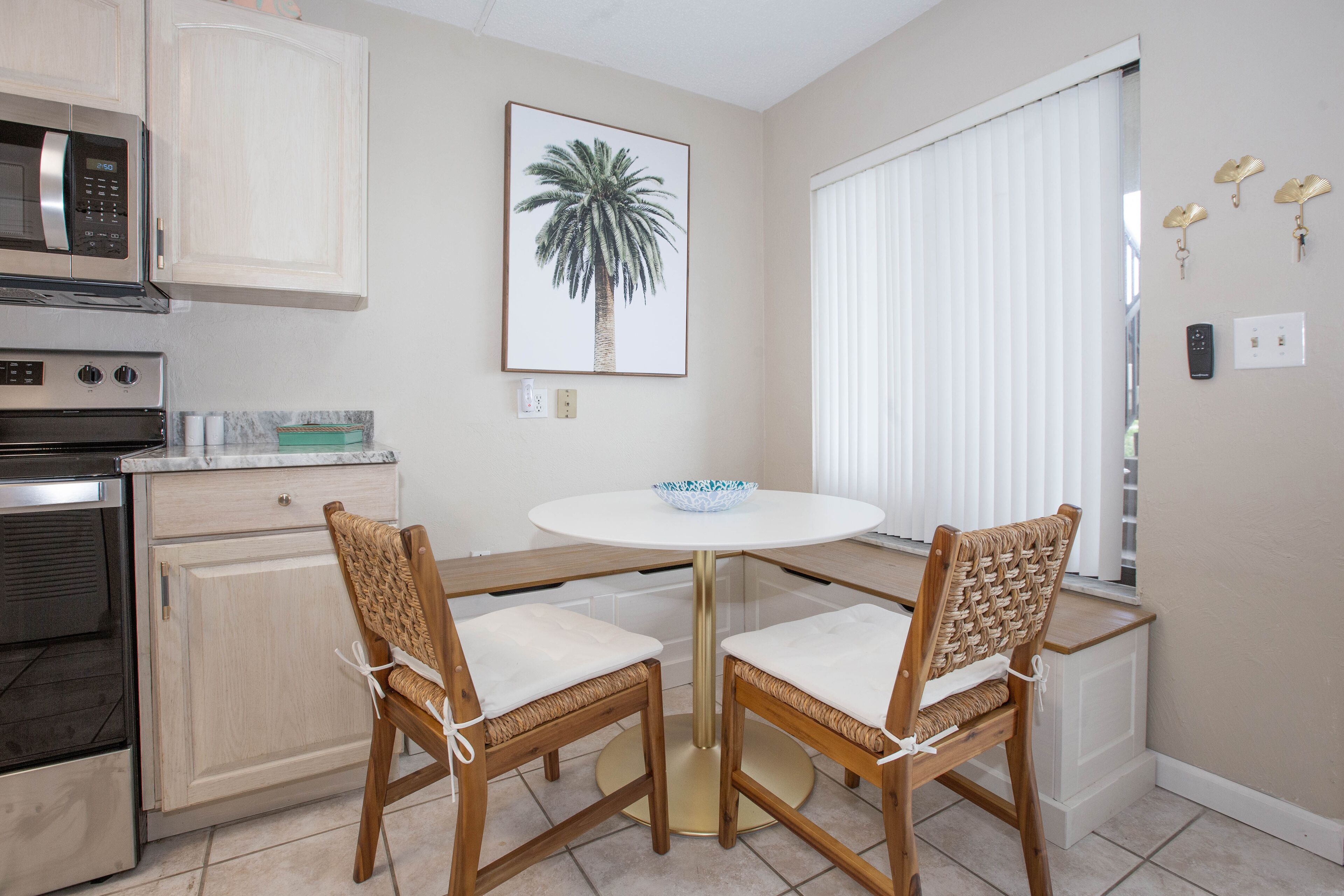 A kitchen with a microwave, oven, and cabinets. There is a framed picture of a palm tree on the wall and a window with blinds.