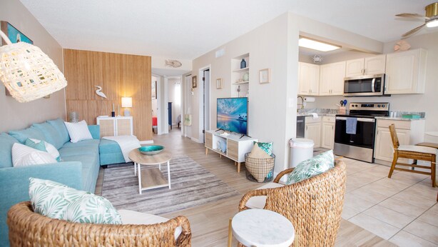 A modern living room with a sofa, wicker chairs, a TV, and a kitchen with white cabinets and appliances.