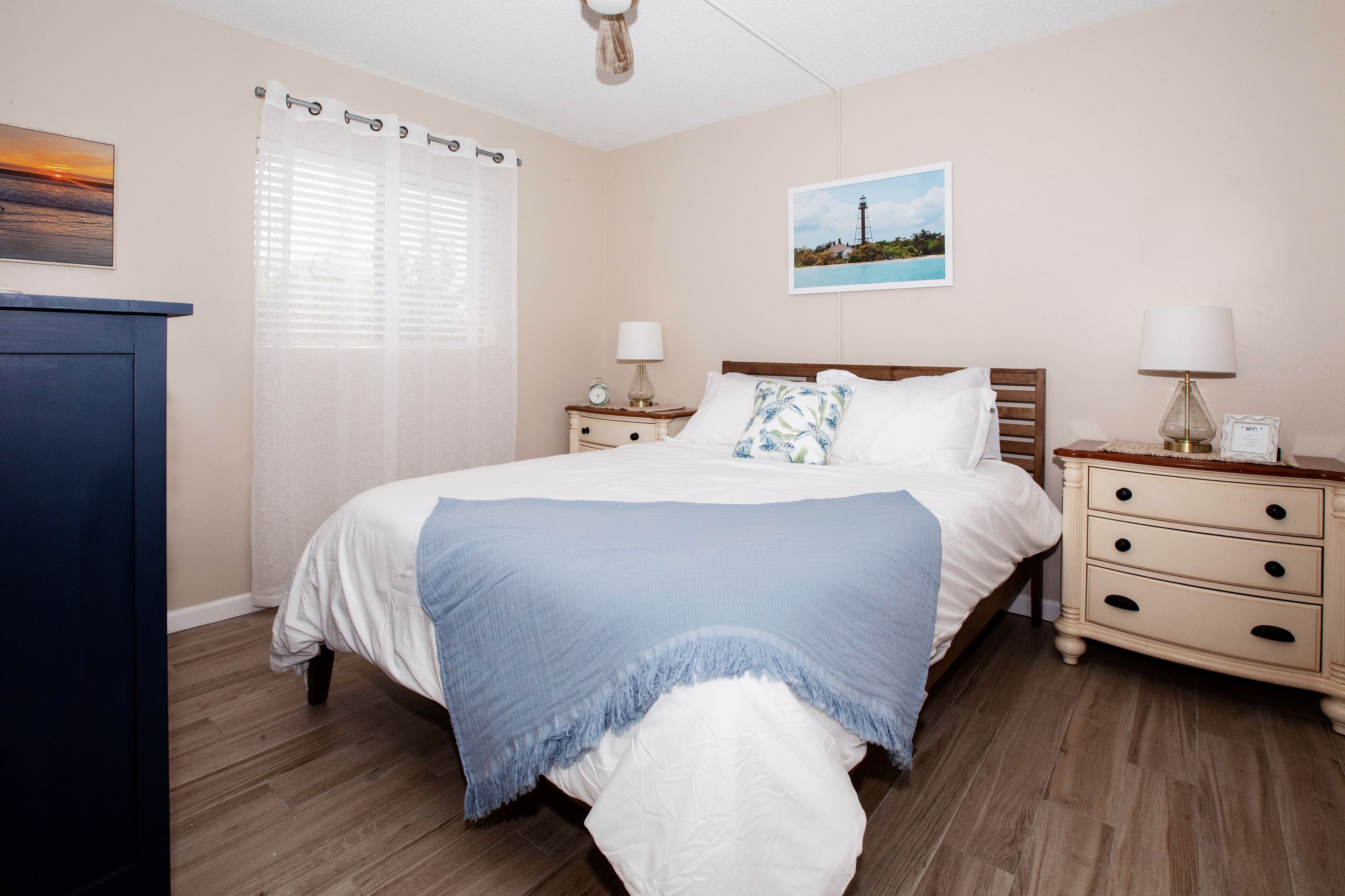 A bedroom with a bed, two bedside tables, a nightstand, a dresser, a window with curtains, and a painting on the wall.