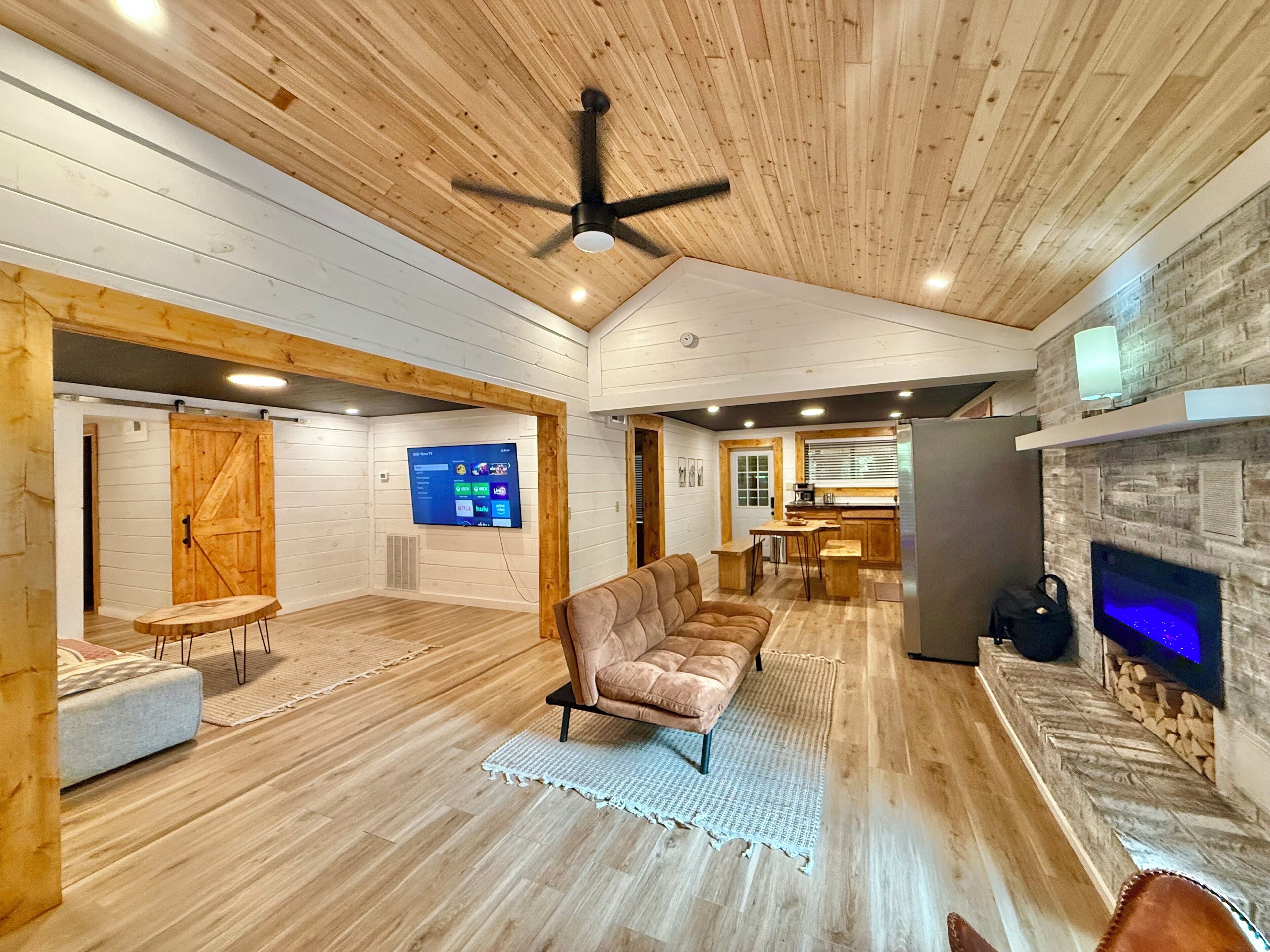 Amplia sala de estar con techo de madera, televisor grande, chimenea y cocina al fondo.