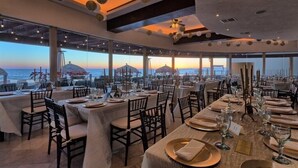 Un restaurant où les tables sont dressées pour le service, de grandes fenêtres offrant une vue sur l’océan et un plafond orné de décorations sphériques suspendues.