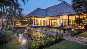 Una casa moderna con un estanque grande, rodeada de plantas tropicales y un camino de piedra.
