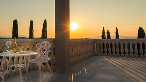 Een terras met een tafel en stoelen, een fles wijn en een uitzicht op de ondergaande zon over een landschap met bomen.