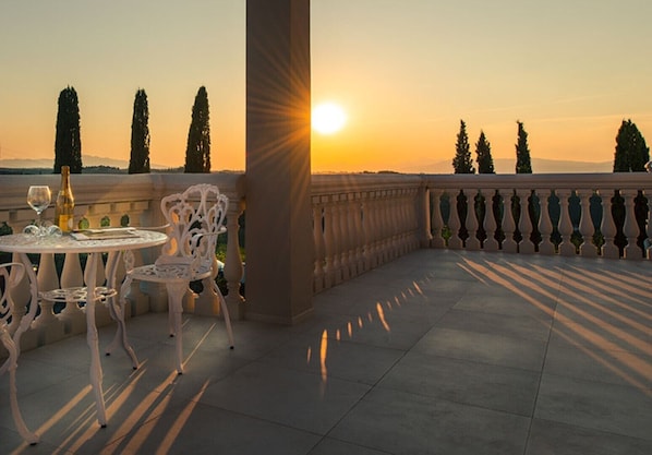 Une terrasse avec une table et des chaises, une bouteille de vin et une vue sur le coucher de soleil, qui se reflète sur un paysage arboré.