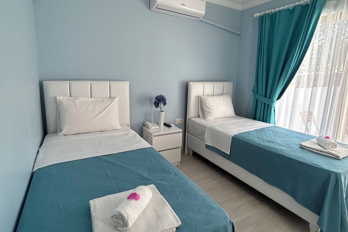 Een hotelkamer met twee bedden, elk met een wit hoofdeinde en turquoise beddengoed. Daartussen bevindt zich een witte nachtkastje, een klein tafeltje met een bloevas, en een raam met turquoise gordijnen.